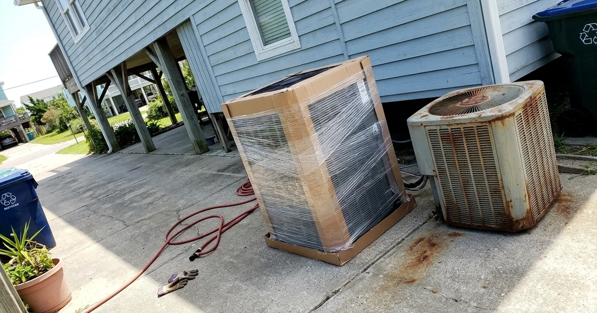 New outdoor AC unit in packaging in a driveway beside the old unit