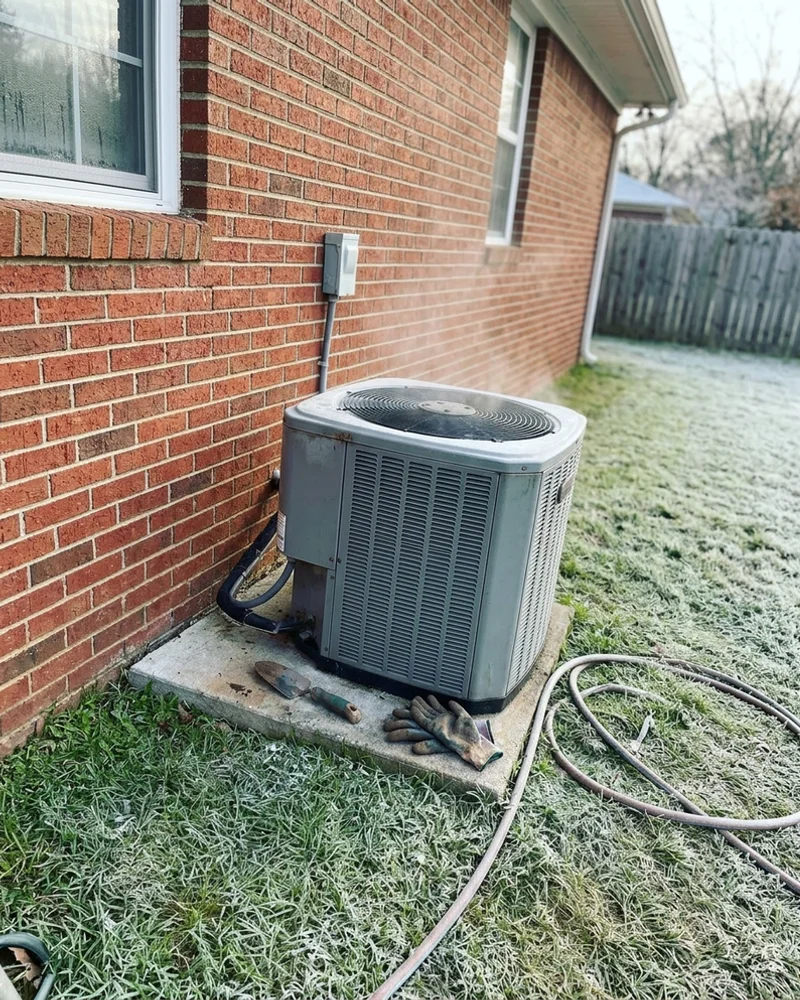 Outdoor heat pump beside a brick home on a chilly winter morning with light frost on the grass